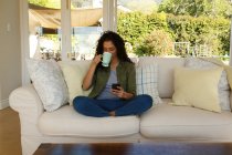 Змішана расова жінка п'є каву за допомогою смартфона, що сидить на дивані у вітальні. самоізоляція в домашніх умовах під час пандемії коронавірусу 19 . — стокове фото