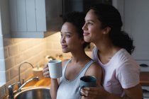 Sorrindo casal lésbico de raça mista bebendo café e abraçando na cozinha. auto isolamento tempo de qualidade em casa juntos durante coronavírus covid 19 pandemia. — Fotografia de Stock