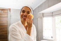 Mujer de raza mixta mirando en espejo cara de limpieza en el baño. autoaislamiento en el hogar durante la pandemia de coronavirus covid 19. - foto de stock