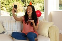Mujer de raza mixta celebrando cumpleaños teniendo chat de vídeo en el teléfono inteligente. llevando sombrero de fiesta y sosteniendo panecillo con vela. autoaislamiento en el hogar durante la pandemia de coronavirus covid 19. - foto de stock