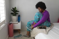 Mujer afroamericana poniéndose ropa deportiva en el dormitorio. autoaislamiento en casa durante la pandemia del coronavirus covid 19. - foto de stock
