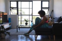 Mujer afroamericana sentada en el sofá poniéndose zapatos deportivos. autoaislamiento en casa durante la pandemia del coronavirus covid 19. - foto de stock