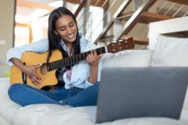 Smiling mixed race woman sitting on couch playing guitar at home. self isolation during covid 19 coronavirus pandemic. — Stock Photo