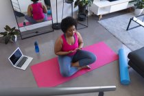 Donna afroamericana che fa meditazione yoga seduta su un tappeto indossando vestiti sportivi. computer portatile in background. auto isolamento fitness benessere tecnologia a casa durante coronavirus covid 19 pandemia. — Foto stock