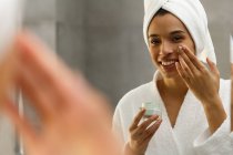 Mujer de raza mixta mirando en espejo aplicando crema facial en el baño. autoaislamiento en el hogar durante la pandemia de coronavirus covid 19. - foto de stock