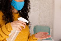 Femme de race mixte portant un masque facial désinfectant les mains avec du spray. portrait d'une femme métisse aux cheveux longs portant un masque facial. auto-isolement à la maison pendant la pandémie de coronavirus covid 19. — Photo de stock