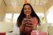 Gemischte Rasse Frau feiert Geburtstag mit Party-Hut mit Muffin mit Kerze darauf. Selbstisolation zu Hause während der Covid 19 Coronavirus-Pandemie. — Stockfoto