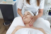 Mujer caucásica tumbada mientras esteticista le da un facial. cliente disfrutando del tratamiento en un salón de belleza. - foto de stock