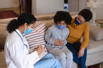 Mixed race mother and daughter talking to mixed race female doctor sitting on couch. self isolation at home together during coronavirus covid 19 pandemic. — Stock Photo