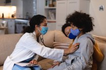 Mixed race girl wearing face mask being examined by mixed race female doctor sitting on couch. self isolation at home together during coronavirus covid 19 pandemic. — Stock Photo