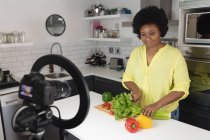 Vlogger afroamericana grabando un video en la cocina. picar verduras. auto aislamiento tecnología comunicación en el hogar durante coronavirus covid 19 pandemia. - foto de stock