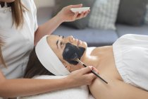 Kaukasische Frau lehnt sich zurück, während Kosmetikerin eine Gesichtsmaske aufträgt. Kundin genießt Behandlung im Schönheitssalon. — Stockfoto