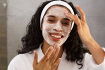 Retrato de mujer de raza mixta aplicando crema facial en el baño. autoaislamiento en el hogar durante la pandemia de coronavirus covid 19. - foto de stock