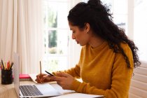 Змішана расова жінка використовує смартфон і ноутбук, сидячи за письмовим столом. самоізоляція в домашніх умовах під час пандемії коронавірусу 19 . — стокове фото