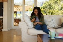Смешанная расовая женщина с помощью смартфона сидит на диване в гостиной. самоизоляция в домашних условиях во время пандемии 19 коронавируса. — стоковое фото