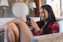 Sorridente donna di razza mista seduta sul divano utilizzando lo smartphone a casa. autoisolamento durante la pandemia di covid 19 coronavirus. — Foto stock