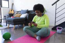 African american woman sitting on exercise mat using smartphone. self isolation fitness technology communication at home during coronavirus covid 19 pandemic. — Stock Photo