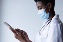 Змішана раса жіночий лікар носив маску обличчя стоячи і використовуючи цифрові таблетки. Гігієна працівників медичного обслуговування під час коронавірусу (19 пандемії). — стокове фото