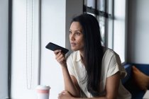 Mujer de negocios de raza mixta de pie junto a la ventana utilizando el teléfono inteligente en la oficina moderna. empresa moderna oficina lugar de trabajo tecnología. - foto de stock
