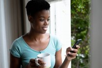 Портрет афро-американської жінки, яка тримає чашку кави, використовуючи смартфон вдома. Залишатися вдома в ізоляції в карантині. — стокове фото