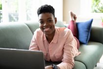 Porträt einer Afroamerikanerin, die mit ihrem Laptop von zu Hause aus auf dem Sofa liegt. Blickt in die Kamera und lächelt. Während der Quarantäne zu Hause bleiben und sich selbst isolieren. — Stockfoto