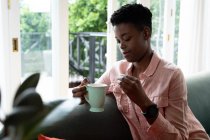 Uma afro-americana sentada no sofá a beber café. ficar em casa em auto-isolamento durante o confinamento de quarentena. — Fotografia de Stock