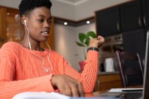 Femme afro-américaine portant des écouteurs faire appel vidéo à l'aide d'un ordinateur portable dans la cuisine. rester à la maison en isolement personnel pendant le confinement en quarantaine. — Photo de stock