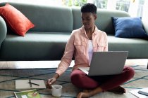 Femme afro-américaine assise sur le sol en utilisant un ordinateur portable buvant du café à la maison. rester à la maison en isolement personnel pendant le confinement en quarantaine. — Photo de stock