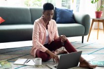 Africano americano mulher sentada no chão usando laptop beber café trabalhando em casa. ficar em casa em auto-isolamento durante o confinamento de quarentena. — Fotografia de Stock