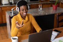 Femme afro-américaine portant des écouteurs faire appel vidéo à l'aide d'un ordinateur portable dans la cuisine. rester à la maison en isolement personnel pendant le confinement en quarantaine. — Photo de stock