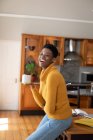 Mujer afroamericana de pie en la cocina tomando café mirando a la cámara y sonriendo. permanecer en casa en aislamiento durante el bloqueo de cuarentena. - foto de stock
