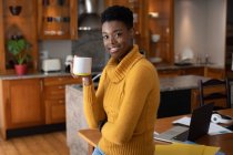 Femme afro-américaine debout dans la cuisine buvant du café en regardant la caméra et souriant. rester à la maison en isolement personnel pendant le confinement en quarantaine. — Photo de stock
