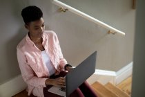 Mujer afroamericana sentada en escaleras usando laptop trabajando desde casa. permanecer en casa en aislamiento durante el bloqueo de cuarentena. - foto de stock