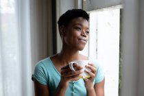Portrait of african american woman holding coffee cup looking out of the window at home. staying at home in self isolation in quarantine lockdown — Stock Photo