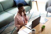 Mulher afro-americana sentada no chão usando laptop falando no smartphone trabalhando em casa. ficar em casa em auto-isolamento durante o confinamento de quarentena. — Fotografia de Stock