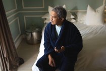 Hombre afroamericano mayor sentado en una cama usando un teléfono inteligente en un dormitorio. estilo de vida de jubilación en aislamiento durante el coronavirus covid 19 pandemia. - foto de stock