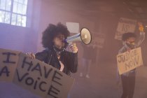 Mujer de raza mixta gritando en megáfono sosteniendo un cartel de protesta. con manifestantes en el fondo. género fluido lgbt identidad concepto de igualdad racial. - foto de stock