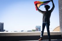 Змішаний чоловік, що стоїть на даху, тримає веселковий прапор. гендерна рідина lgbt ідентифікація концепція расової рівності . — стокове фото