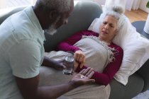 Femme caucasienne âgée couchée malade sur le canapé homme passant la médecine et l'eau dans le salon. rester à la maison en isolement personnel pendant le confinement en quarantaine. — Photo de stock