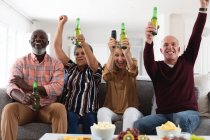 Senior caucasian and african american couples sitting on couch watching game drinking beer at home. senior retirement lifestyle friends socializing. — Stock Photo