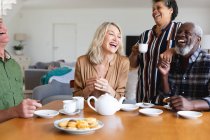 Senior caucasian and african american couples sitting by table drinking tea at home. senior retirement lifestyle friends socializing. — Stock Photo