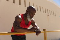 Ajuste o homem americano africano sênior que exercita-se usando smartwatch que inclina-se na cerca no sol. tecnologia de aposentadoria saudável comunicação ao ar livre fitness lifestyle. — Fotografia de Stock