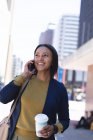 Mujer afroamericana con taza de café hablando en smartphone en la calle. estilo de vida durante la pandemia de coronavirus covid 19. - foto de stock