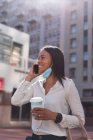 Femme afro-américaine avec masque facial abaissé parlant sur smartphone dans la rue. mode de vie concept de vie pendant coronavirus covid 19 pandémie. — Photo de stock