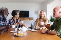 Старшие кавказские и африканские американские пары сидят за столом и пьют чай дома. Друзья на пенсии общаются. — стоковое фото