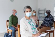 Разнообразная группа пожилых людей в масках для лица разговаривает во время групповой терапии дома. здоровье гигиены благополучие в доме престарелых во время коронавирусной ковиды 19 пандемии. — стоковое фото