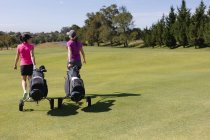 Дві бігові жінки, що йдуть через поле для гольфу, розмовляють, тягнучи сумки для гольфу на колесах. спорт дозвілля хобі гольф здоровий спосіб життя на відкритому повітрі . — стокове фото