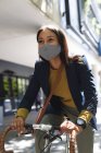 African american woman wearing face mask riding bicycle on the street. lifestyle living during coronavirus covid 19 pandemic. — Stock Photo