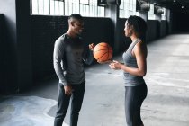 Homem e mulher afro-americanos de pé em um prédio urbano vazio e jogando basquete. fitness urbano estilo de vida saudável. — Fotografia de Stock