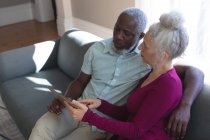 Senioren-Mixed-Race-Paar sitzt auf der Couch und schaut gemeinsam auf das digitale Tablet im Wohnzimmer. Während der Quarantäne zu Hause bleiben und sich selbst isolieren. — Stockfoto